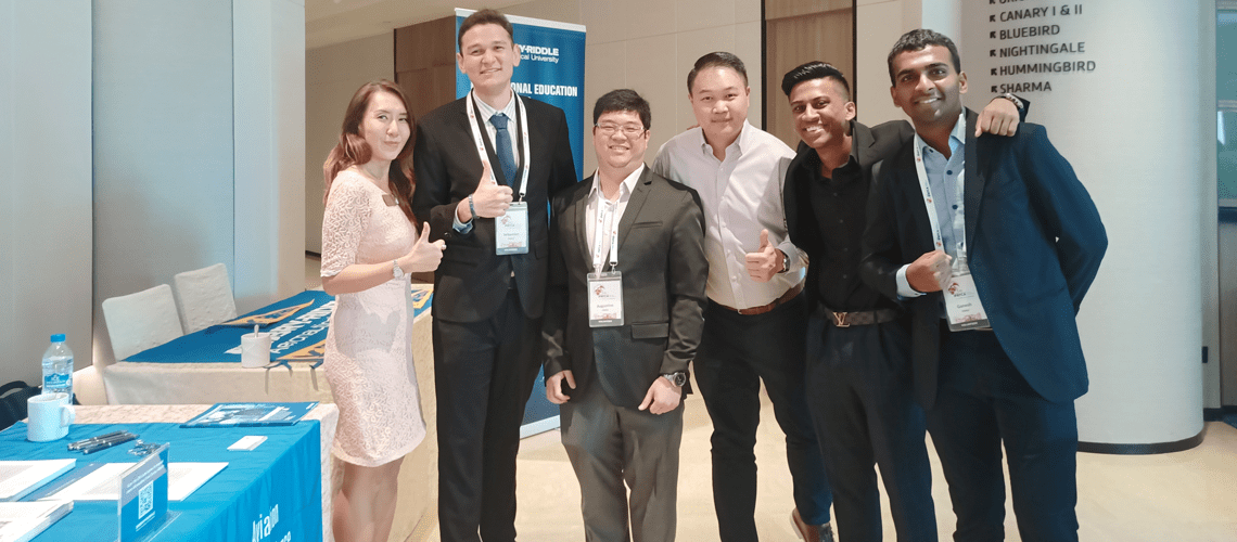 Six students in business wear pose near an expo table with Embry-Riddle banners and tablecloth. Three of the students are giving a thumbs up.